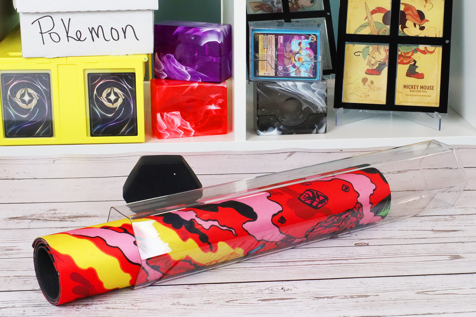 A rolled red, yellow, and black stitched playmat inside a clear hexagon-shaped BCW Large Playmat Tube, displayed on a wooden table in front of a shelving unit with trading card storage boxes, magnetic card holders, and framed artwork.